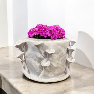 May include: A gray, textured ceramic planter with mushroom-shaped accents, filled with vibrant pink flowers. The planter sits on a marble countertop, creating a contrast of textures and colors. A unique decorative piece for home decor.