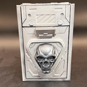 May include: A rectangular silver-colored storage box featuring a three-dimensional skull emblem centered on the front panel. The box has a textured, industrial-style surface with geometric molded details, including a latch mechanism above the skull.