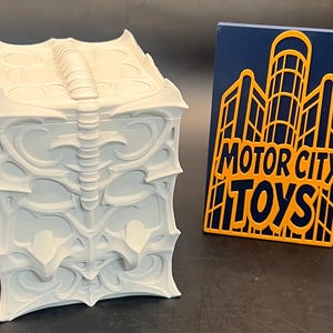 May include: A light gray, ornate box with a textured surface and a hinged lid. The box features a detailed, stylized design. Next to the box is a blue sign with the words "MOTOR CITY TOYS" in yellow.