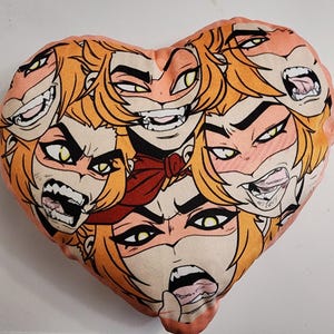 May include: A heart-shaped pillow with a repeating pattern of cartoon faces with yellow eyes, red lips, and orange hair. The faces are all different expressions, including smiling, frowning, and sticking out their tongues.