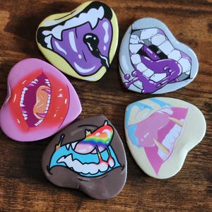 Pode incluir: Seis pinos em forma de coração com diferentes desenhos de boca coloridos. Cada pino apresenta um estilo de boca diferente com dentes, língua e líquido pingando. Os pinos são uma mistura de rosa, roxo, amarelo, azul, marrom e branco.