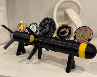 May include: A black and yellow 3D-printed missile-shaped coin display stand holding several collectible coins. The stand has fins and support legs. The coins have various designs and colors, including gold, black, and red.