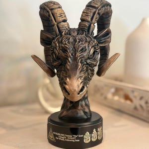 May include: A bronze-colored ram head sculpture with large curved horns, mounted on a black base. The base has text and emblems, including "Tommy The Goat" and "Fair Winds, Following Seas". The sculpture is detailed and textured.