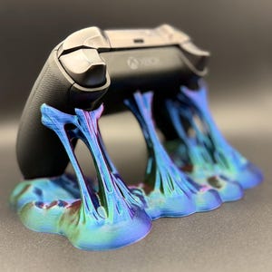 May include: A black Xbox controller is displayed on a iridescent blue and green 3D printed stand. The stand has a unique, organic design.