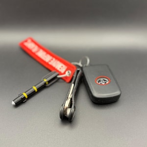 May include: A black car key with a red logo, a black leather key chain, and a red and white keychain with the text "VERIFIED" on it.