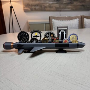 Peut inclure: Un présentoir gris en forme de missile contenant diverses pièces de monnaie militaires et un petit panneau noir avec le texte "Match Arrows". Le présentoir a une pointe rouge et de petites ailes. Les pièces présentent différents emblèmes et motifs.