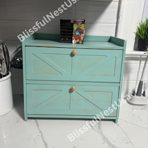 May include: A two-drawer turquoise bread box with wooden knobs. The doors have a farmhouse style design. A small decorative item is on top of the bread box.