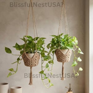 May include: Two hanging planters featuring light brown crocheted holders with tassels, each containing a green pothos plant. The text BlissfulNestUSA is visible in the image. The planters are suspended from black hooks against a neutral wall.