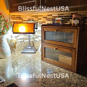 May include: A two-tiered wooden bread box with glass doors. The bread box has a dark wood finish and brass hardware. The top tier has a small box on top of it with a jar of snacks. The bread box is sitting on a granite countertop in a kitchen.