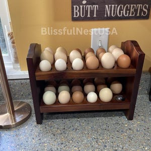 May include: A two-tiered wooden egg rack filled with various colored eggs. The eggs range in color from white to brown. A sign above the rack reads "farm fresh BUTT NUGGETS".