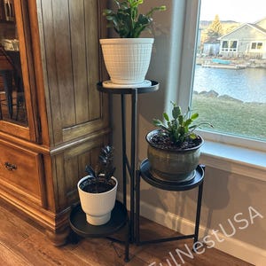 May include: A black metal three-tier plant stand holding three potted plants. The top plant is in a white textured pot, the middle plant is in a green ribbed pot, and the bottom plant is in a white ribbed pot. Text reads BlissFulNestUSA.