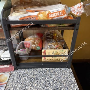 May include: A two-tier storage rack with a dark metal frame and wood-grain shelves. The top shelf holds a package of Thomas' Original English Muffins. The bottom shelf contains a plastic container, a loaf of wheat bread, and a box of Costa Rica coffee.