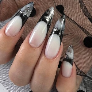 May include: Close-up of a hand with long, oval-shaped acrylic nails. The nails feature a white base with a black tip and silver foil accents. The nails are set against a black mesh fabric.