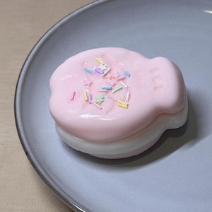 May include: A pink and white dessert, possibly a pastry or soap, sits on a gray plate. The top layer is a light pink with colorful sprinkles. The bottom layer is white. The dessert is round with a wavy edge.