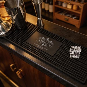 May include: A rectangular black rubber bar mat with a textured nub surface. The center features a smooth black panel printed with the text "Williams's Bar" and "Good Friends, Good Drinks, Great Times" in white, surrounded by decorative scrollwork.