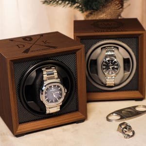 May include: Two wooden watch winders with a black and gray interior. The watch winders have a clear glass top and are engraved with initials and a date. One winder has a silver watch inside.
