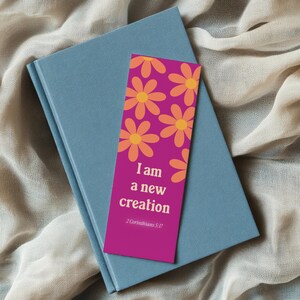 May include: A blue book with a bookmark. The bookmark is magenta with orange and yellow flower graphics. The bookmark has the text "I am a new creation" and "2 Corinthians 5:17".