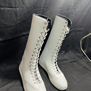 May include: A pair of white leather lace-up boots with black laces. The boots are tall and have a rounded toe. The boots are on a black background.