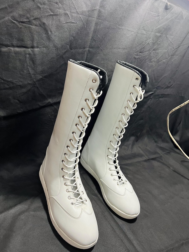 May include: A pair of white leather lace-up boots with black laces. The boots are tall and have a rounded toe. The boots are on a black background.