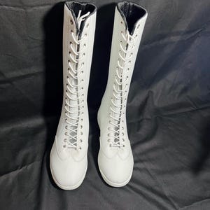 May include: A pair of white leather lace-up boots with black trim around the top of the boot.