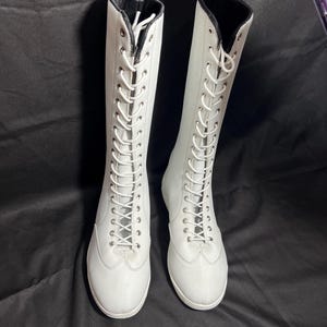 May include: A pair of white lace-up boots with black trim around the top of the boot.