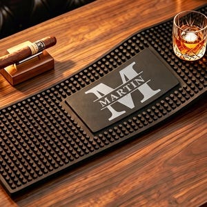 May include: A rectangular black rubber bar mat with a textured surface featuring a central panel printed with a large letter M and the name MARTIN. The mat is positioned on a wooden surface alongside a glass of amber liquid and two small gold jiggers.