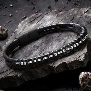 Puede incluir: Pulsera de cuero negro con detalles plateados y cierre magnético. La pulsera se muestra sobre una superficie de piedra gris oscura, resaltando su diseño moderno. Un accesorio para el uso diario.