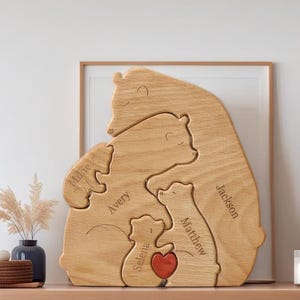 Pode incluir: Quebra-cabeça de família de ursos de madeira com cinco figuras de ursos. Cada urso tem um tamanho diferente e um nome gravado: Millie, Avery, Selena, Matthew e Jackson. Um coração vermelho está no centro. O quebra-cabeça está em uma prateleira de madeira.