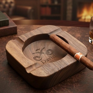 May include: A square wooden ashtray with a cigar resting in it. The ashtray has a carved design and the name "David" engraved on it. A glass of amber liquid sits nearby. The ashtray is on a brown surface.