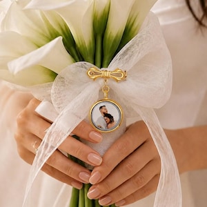 May include: A bouquet of white calla lilies tied with a white sheer ribbon. A gold-toned metal charm featuring a circular photograph of a couple is attached to the ribbon, positioned centrally against the stems of the flowers.