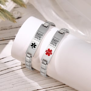 Può includere: Due braccialetti di allerta medica in acciaio inossidabile. Uno presenta un simbolo medico nero, l'altro rosso. Entrambi hanno un codice QR e il testo "SCAN ME" e "HEALTH INFO".