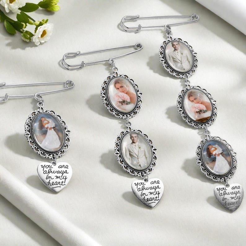 May include: Three silver-toned safety pin brooches featuring oval photo frames with wedding portraits. Each brooch includes a dangling heart-shaped charm engraved with the text, "you are always in my heart!" The frames have ornate, scalloped edges.
