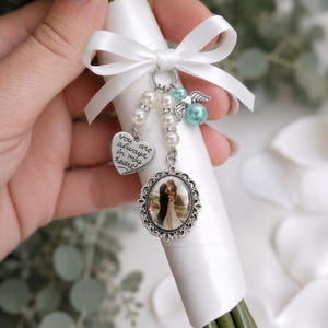 May include: A white satin ribbon-wrapped bouquet handle charm featuring a silver heart pendant engraved with the text 'you are always in my heart', a silver oval photo frame pendant, white faux pearls, and a turquoise bead with silver angel wings.
