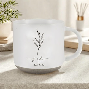 May include: White ceramic mug with a grey base. The mug features a black botanical illustration and the inscription "j + h" with the date "02.12.25". The mug has a curved handle.