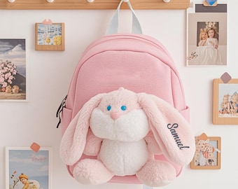 May include: A pink backpack with a plush bunny attached. The bunny has long ears, blue eyes, and the name "Samuel" printed on one ear. The backpack hangs from a wooden rack with other framed pictures.