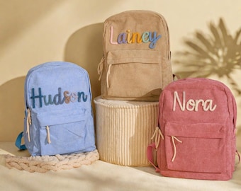 May include: Four corduroy backpacks in blue, beige, and pink, each with a name embroidered on the front. The names are "Hudson", "Lainey", and "Nora". The backpacks have a front pocket and side pockets.