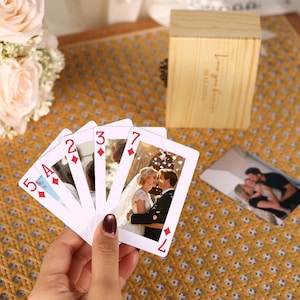 Puede incluir: Una mano sostiene un abanico de cinco cartas de juego personalizadas con diamantes y fotos de una pareja. Las cartas muestran los números 5, A, 2, 3 y 7. Una caja de madera clara con el texto Love you forever 10.12.2024 es visible cerca.