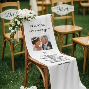 May include: White fabric chair sash with the text "IN LOVING memory" and a photo of a couple. The sash also includes the text "WE ARE IN HEAVEN...WE WILL BE THERE." A bouquet of white flowers is attached to the chair. The chair is part of a wedding ceremony.