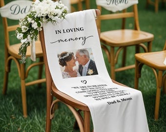 May include: White fabric chair sash with the text "IN LOVING memory" and a photo of a couple. The sash also includes the text "WE ARE IN HEAVEN...WE WILL BE THERE." A bouquet of white flowers is attached to the chair. The chair is part of a wedding ceremony.