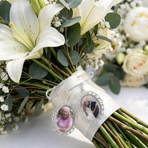 May include: A silver-toned metal brooch pin featuring two oval-shaped photo frames with ornate filigree borders. The pin is attached to a white ribbon wrapped around a bouquet of white lilies, eucalyptus leaves, and baby's breath flowers.