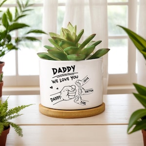 Peut inclure: Pot de fleurs en céramique blanche avec une plante succulente verte. Le pot porte l'inscription "DADDY WE LOVE YOU" avec des illustrations de poings et des noms. Il repose sur un dessous de plat rond en bambou brun clair. Le fond comprend des plantes et une fenêtre.
