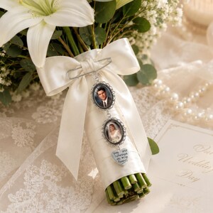 May include: A silver-toned memorial charm set featuring two oval photo frames and a heart-shaped pendant with the text "you are always in my heart" attached to a white satin ribbon bow wrapped around a bouquet of white lilies and greenery.