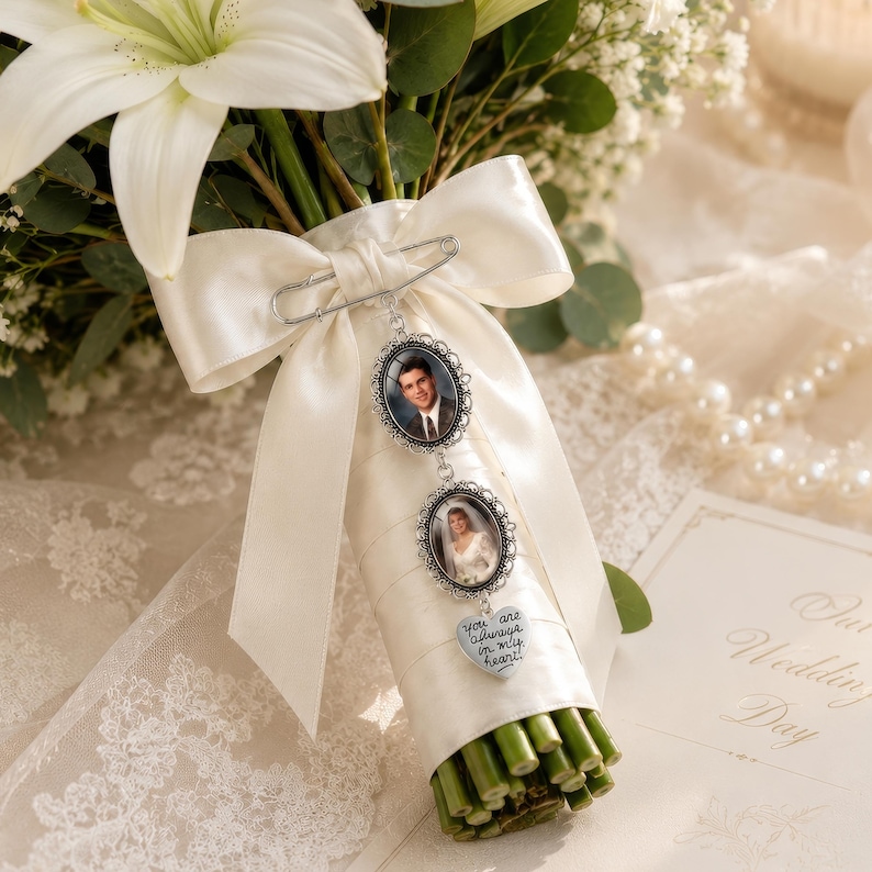 May include: A silver-toned memorial charm set featuring two oval photo frames and a heart-shaped pendant with the text "you are always in my heart" attached to a white satin ribbon bow wrapped around a bouquet of white lilies and greenery.