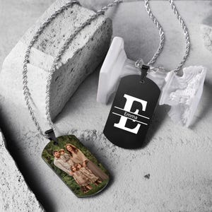 Könnte beinhalten: Zwei personalisierte Dog-Tag-Halsketten mit silbernen Ketten. Ein Anhänger zeigt ein Familienfoto, der andere einen schwarzen Anhänger mit dem Buchstaben "E" und dem Namen "Emma" in Weiß. Die Anhänger werden auf einer strukturierten Oberfläche präsentiert.