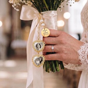 May include: A gold-toned wedding bouquet charm featuring two heart-shaped frames with black and white photographs, suspended from a cream satin ribbon. A round gold disc is engraved with the text Brittany & Michael 06-11-26, accented by two white pearls.