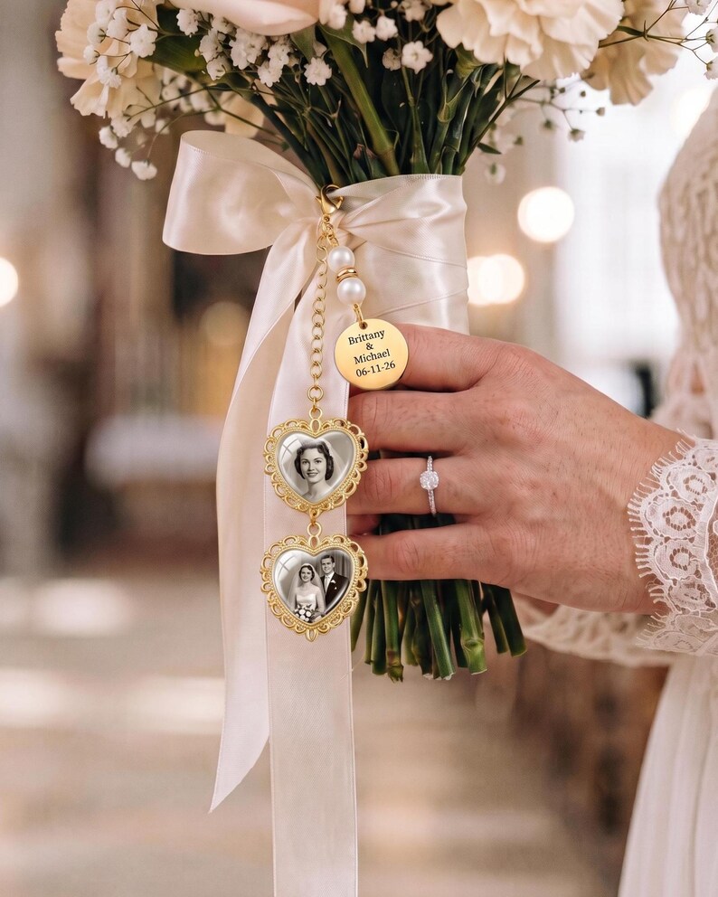 May include: A gold-toned wedding bouquet charm featuring two heart-shaped frames with black and white photographs, suspended from a cream satin ribbon. A round gold disc is engraved with the text Brittany & Michael 06-11-26, accented by two white pearls.