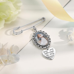 May include: A silver-toned safety pin brooch featuring an oval pendant with a decorative filigree frame. The pendant displays a photograph of a person in a white dress. A heart-shaped charm hangs below, engraved with the text, You are always in my heart.