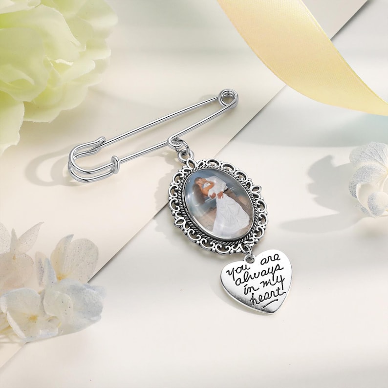 May include: A silver-toned safety pin brooch featuring an oval pendant with a decorative filigree frame. The pendant displays a photograph of a person in a white dress. A heart-shaped charm hangs below, engraved with the text, You are always in my heart.