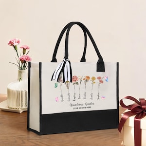 Puede incluir: Una bolsa de lona blanca con asas negras y fondo negro. La bolsa presenta un diseño floral con mariposas y el texto "Grandma's Garden Love Grows Here".
