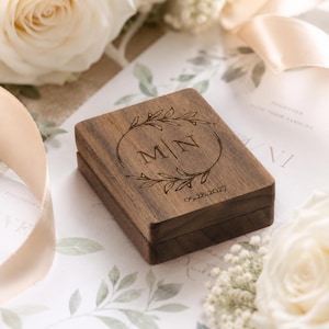 Peut inclure: Une boîte à bagues rectangulaire en bois brun foncé avec un motif gravé au laser sur le couvercle. La gravure présente les lettres M et N séparées par une ligne verticale, entourées d'une couronne de laurier, avec la date 05.28.2027 en dessous.